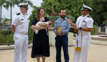 Naval officers stand alongside mother holding a baby and a man holding the Navy quarterdeck bell.