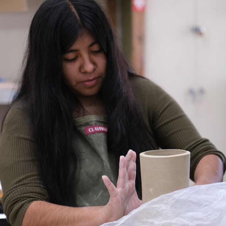 Student shaping a clay cylinder while working at a ceramics studio table.