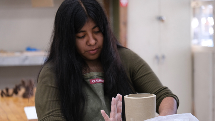 Student shaping a clay cylinder while working at a ceramics studio table.