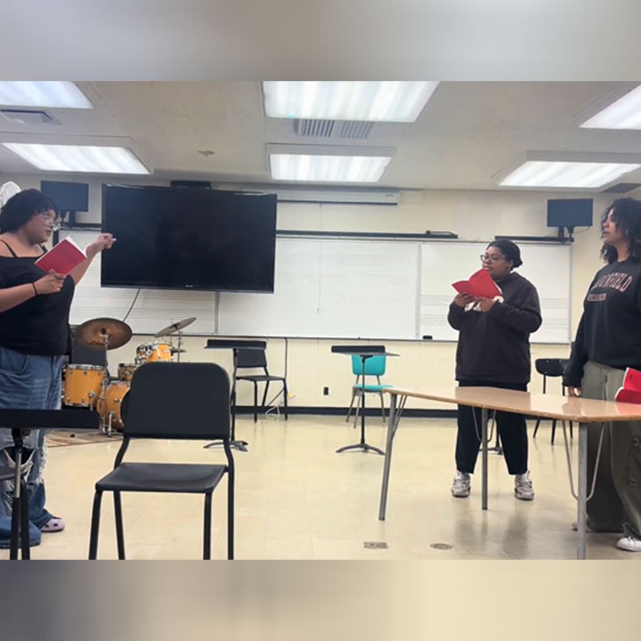 Students rehearsing with scripts in a music classroom, with chairs, instruments and a piano visible.