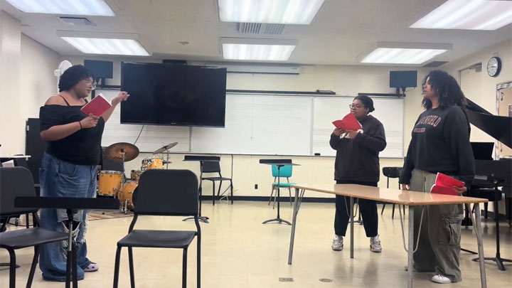 Students rehearsing with scripts in a music classroom, with chairs, instruments and a piano visible.