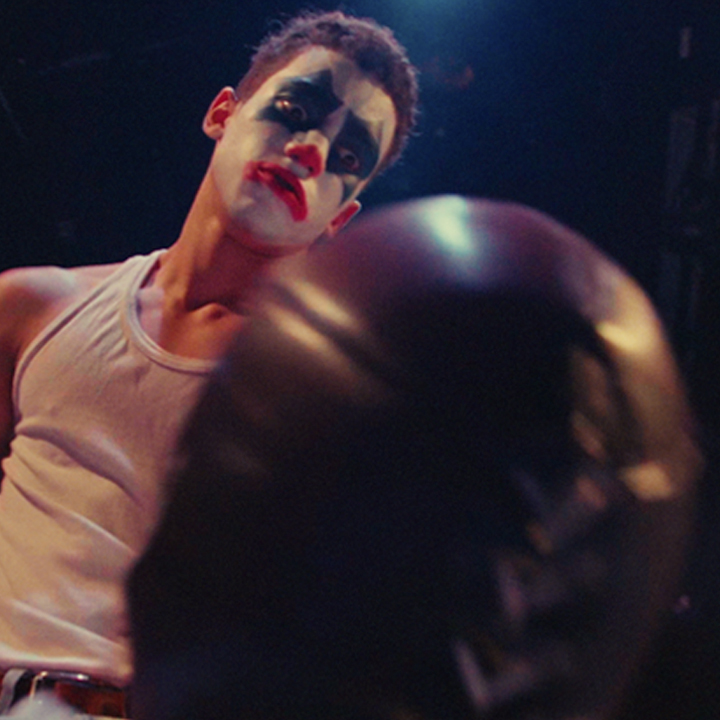 Dramatic film still of a performer in face paint confronting another figure under stage lighting.