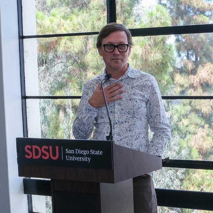 Speaker standing at an SDSU podium giving remarks in front of large windows.