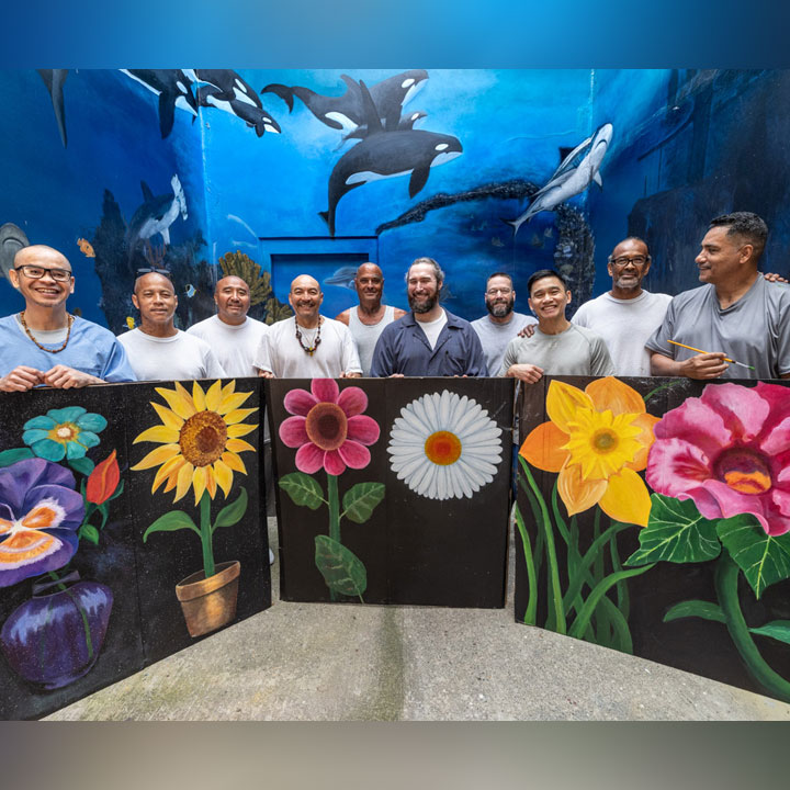 Group of incarcerated men posing with large flower paintings during an art program, with a mural-covered wall behind them.