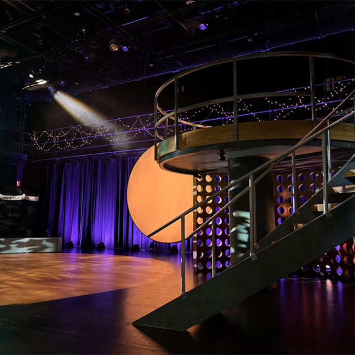  Theater stage with dramatic lighting, spiral staircase set piece, and decorative string lights.